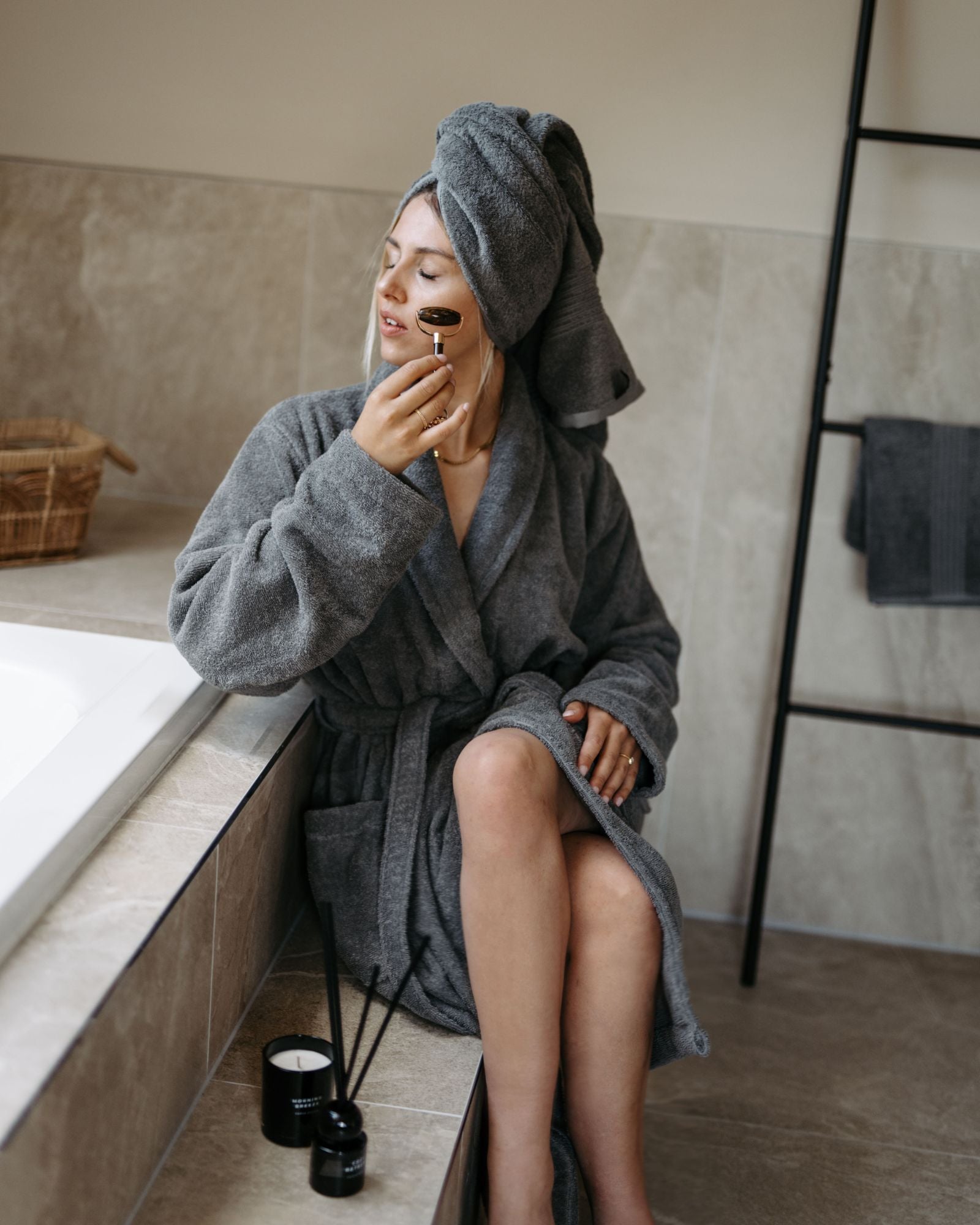 Junge blonde Frau beim Wellness Zuhause mit einem Gesichtsroller, Frottier-Bademantel in grau und Handtuch auf dem Kopf