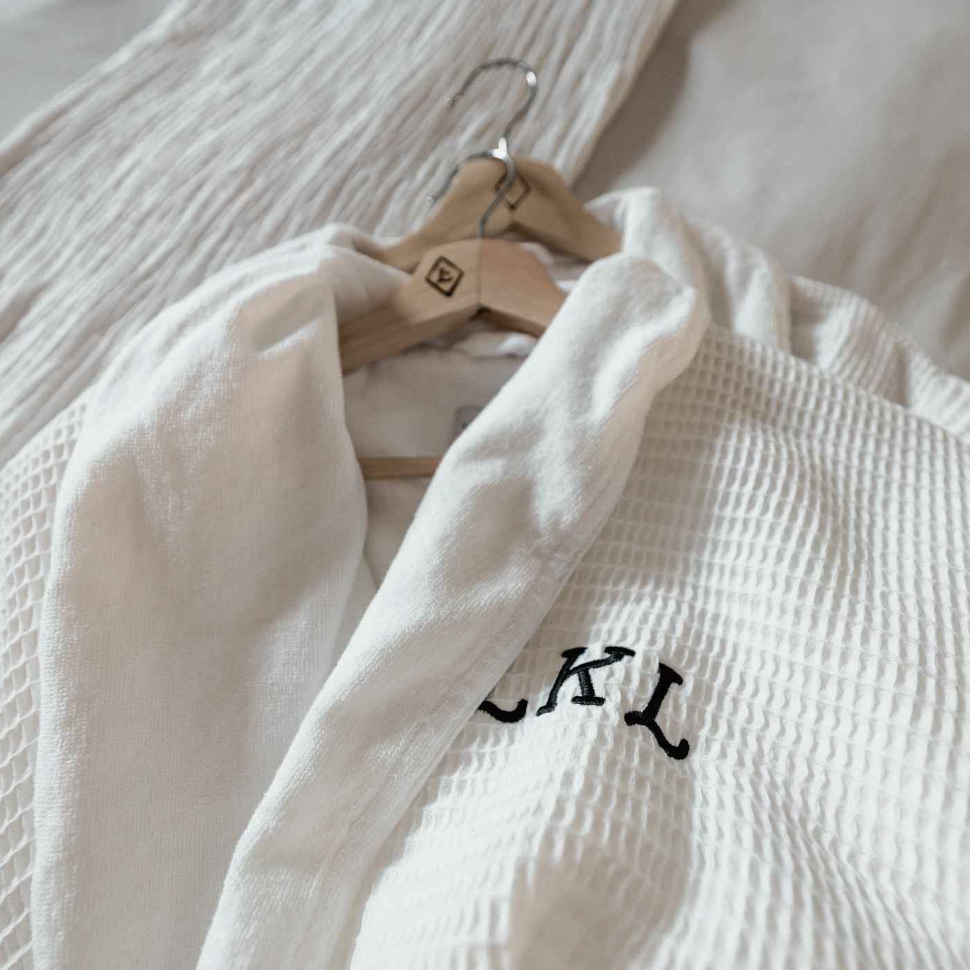 Weißer Bademantel mit feiner Waffelstruktur und personalisierter Stickerei, hängend auf einem Holzkleiderbügel – ideal für Hotels und Spas