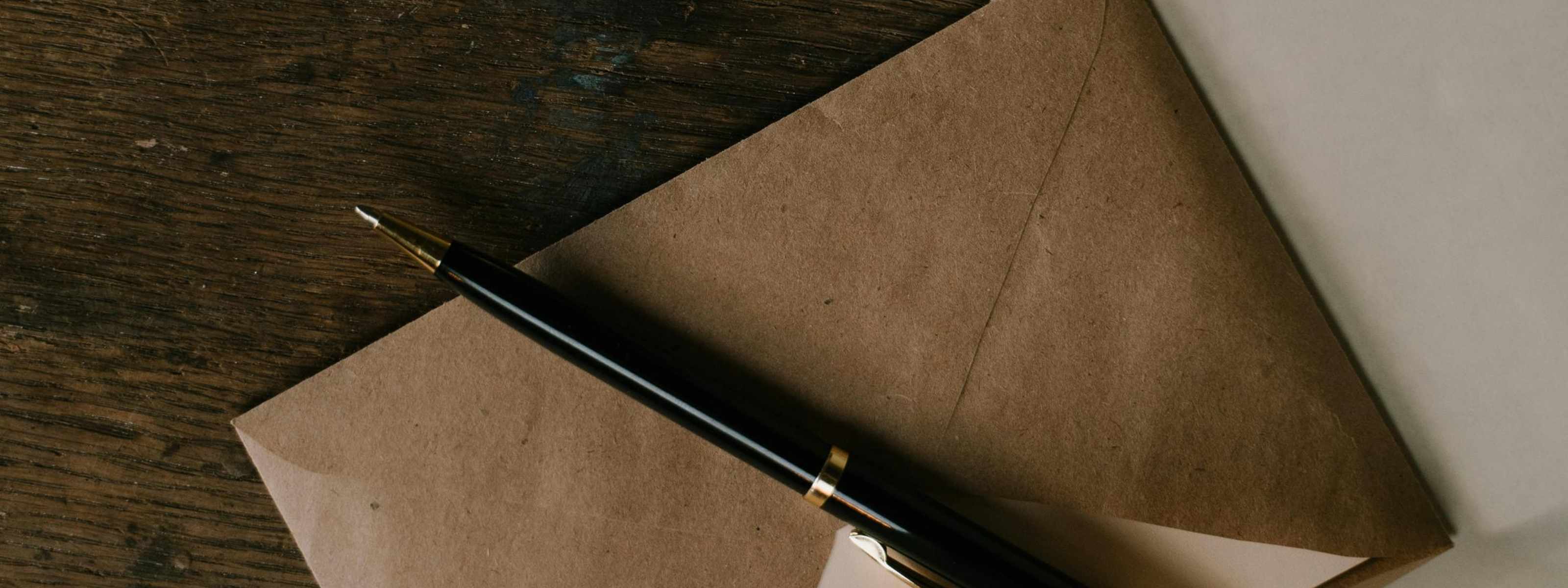 Ein brauner Briefumschlag mit einem schwarzen Füllfederhalter liegt auf einer rustikalen Holzoberfläche, daneben ein weißes Blatt Papier